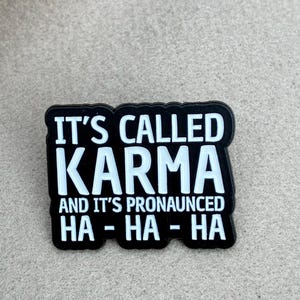 May include: A rectangular black enamel pin with white raised lettering that reads IT'S CALLED KARMA AND IT'S PRONOUNCED HA-HA-HA. The pin features a bold, sans-serif font with a clean, high-contrast design and a smooth, glossy finish.