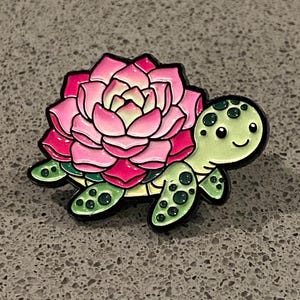 May include: A decorative enamel pin in the shape of a turtle, with a succulent flower shell. The turtle has a green head and flippers with black spots. The flower is pink and white with black outlines.