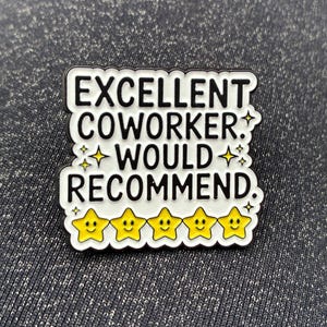 Puede incluir: Un pin de esmalte blanco con texto negro que dice EXCELLENT COWORKER. WOULD RECOMMEND. El diseño presenta cinco estrellas amarillas con caras sonrientes en la parte inferior y pequeños acentos de estrellas amarillas alrededor del texto.