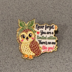 Peut inclure: Une broche en émail doré représentant un hibou brun aux grands yeux portant un bandeau vert à motifs de feuilles. À côté du hibou se trouve un panneau rectangulaire blanc à bordure dorée avec le texte : Never forget: You are a limited edition. There's no one like you!