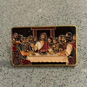 Op de afbeelding: Een rechthoekige emaille speld met een gedetailleerde afbeelding van het Laatste Avondmaal. De scène toont een centrale figuur in wit en rood gewaad aan een lange tafel met twaalf andere figuren. De rand is van goudkleurig metaal.