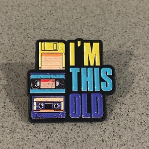 May include: A black enamel pin featuring a yellow floppy disk, a light blue cassette tape, and a dark blue cassette tape stacked vertically. To the right, the text I'M THIS OLD is displayed in yellow, light blue, and dark blue block letters.
