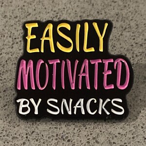 Peut inclure: Épinglette rectangulaire noire avec le texte "EASILY MOTIVATED BY SNACKS". Le texte est en jaune, rose et blanc. L'épinglette est probablement un accessoire de chaussure.