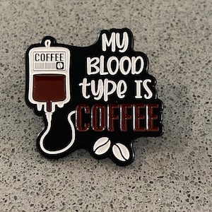 Könnte beinhalten: Schwarzer und weißer Anstecker mit dem Text "MY BLOOD TYPE IS COFFEE". Der Anstecker zeigt einen Kaffee-Infusionsbeutel, Kaffeebohnen und das Wort "COFFEE" in braunen Buchstaben.