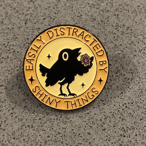May include: A round enamel pin with a pale yellow background and a black border. The centre features a black bird holding a small gold-framed purple gem. The text "EASILY DISTRACTED BY SHINY THINGS" is printed in black along the circular edge.