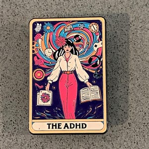 Peut inclure: Un pin's rectangulaire en émail représentant une carte de tarot sur fond bleu foncé. Un personnage central en chemise blanche et pantalon rose est entouré de motifs tourbillonnants colorés, d'icônes comme une horloge, un livre et un casque, avec le texte THE ADHD.