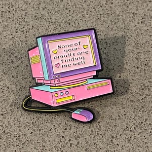 May include: A colourful enamel pin shaped like a vintage desktop computer with a pink and light blue casing. The screen displays the text 'NONE of your emails are finding me well' surrounded by small yellow and pink heart icons, with a mouse attached.