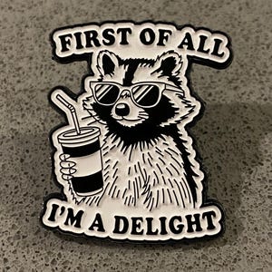 以下が含まれることがあります： サングラスをかけたアライグマが飲み物を持っている、白黒のエナメルピン。ピンには「FIRST OF ALL I'M A DELIGHT」という白い文字が黒い縁取りで書かれています。灰色の背景に配置されています。