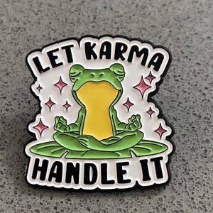 Peut inclure: Épingle en émail représentant une grenouille méditant sur une feuille de nénuphar. La grenouille est verte et jaune, avec le texte "LET KARMA HANDLE IT" en noir et blanc. Étoiles roses en arrière-plan.