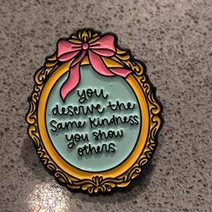 Peut inclure: Une broche en émail de forme ovale avec un cadre doré orné et un nœud rose sur le dessus. Le centre est bleu clair avec le texte noir : you deserve the same kindness you show others. La broche présente un contour noir et une finition lisse.