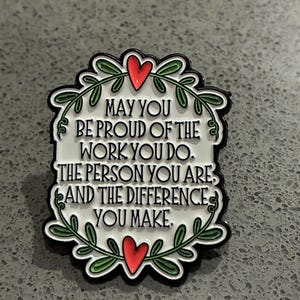 Peut inclure: Une broche en émail blanc avec le texte noir : MAY YOU BE PROUD OF THE WORK YOU DO. THE PERSON YOU ARE, AND THE DIFFERENCE YOU MAKE. La broche est encadrée par une bordure de vigne verte avec deux cœurs rouges au centre en haut et en bas.