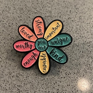 Könnte beinhalten: Eine blumenförmige Emaille-Anstecknadel mit einem türkisfarbenen Zentrum, das "you are" liest, und acht Blütenblättern in Rosa-, Orange- und Türkistönen. Jedes Blütenblatt trägt schwarzen kursiven Text: "loved", "strong", "important", "unique", "kind", "capable", "enough" und "worthy".