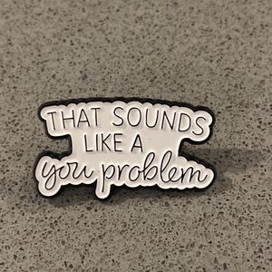 May include: A white and black pin featuring the text "THAT SOUNDS LIKE A you problem" in a mix of block and cursive fonts. The pin has a black border and is set against a speckled grey surface.