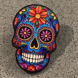 May include: A decorative enamel pin shaped like a human skull, featuring a vibrant blue base colour with intricate, multi-coloured floral patterns in shades of orange, pink, purple, and yellow, complete with a black outline and white teeth.