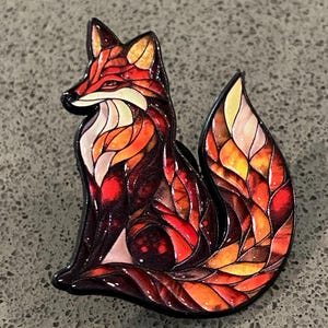 Könnte beinhalten: Eine fuchsförmige Emaille-Anstecknadel mit einem Design im Buntglas-Stil. Der Fuchs ist in leuchtenden Orange-, Rot-, Gelb- und tiefen Burgundertönen gehalten, wobei schwarze Umrandungen jedes Segment trennen und einen Mosaikeffekt erzeugen.