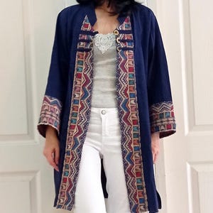 Puede incluir: Una chaqueta larga azul marino con un estampado geométrico colorido en el ribete. La chaqueta tiene un cuello mandarín y mangas largas. La modelo lleva pantalones vaqueros blancos y tacones negros.