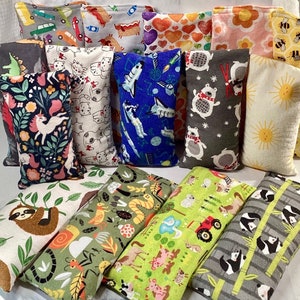 May include: A collection of 14 colorful fabric-covered heat packs with various designs, including animals, space, and nature scenes. The heat packs are rectangular and have a sewn seam down the center.