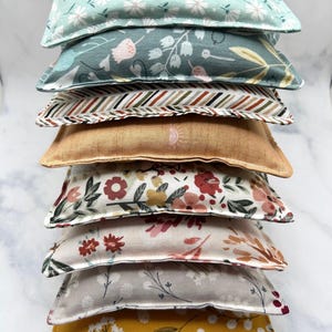 May include: A stack of eight fabric-covered heat packs in various floral and geometric patterns. The heat packs are arranged from largest to smallest, with the top pack being a light blue with a white floral pattern.