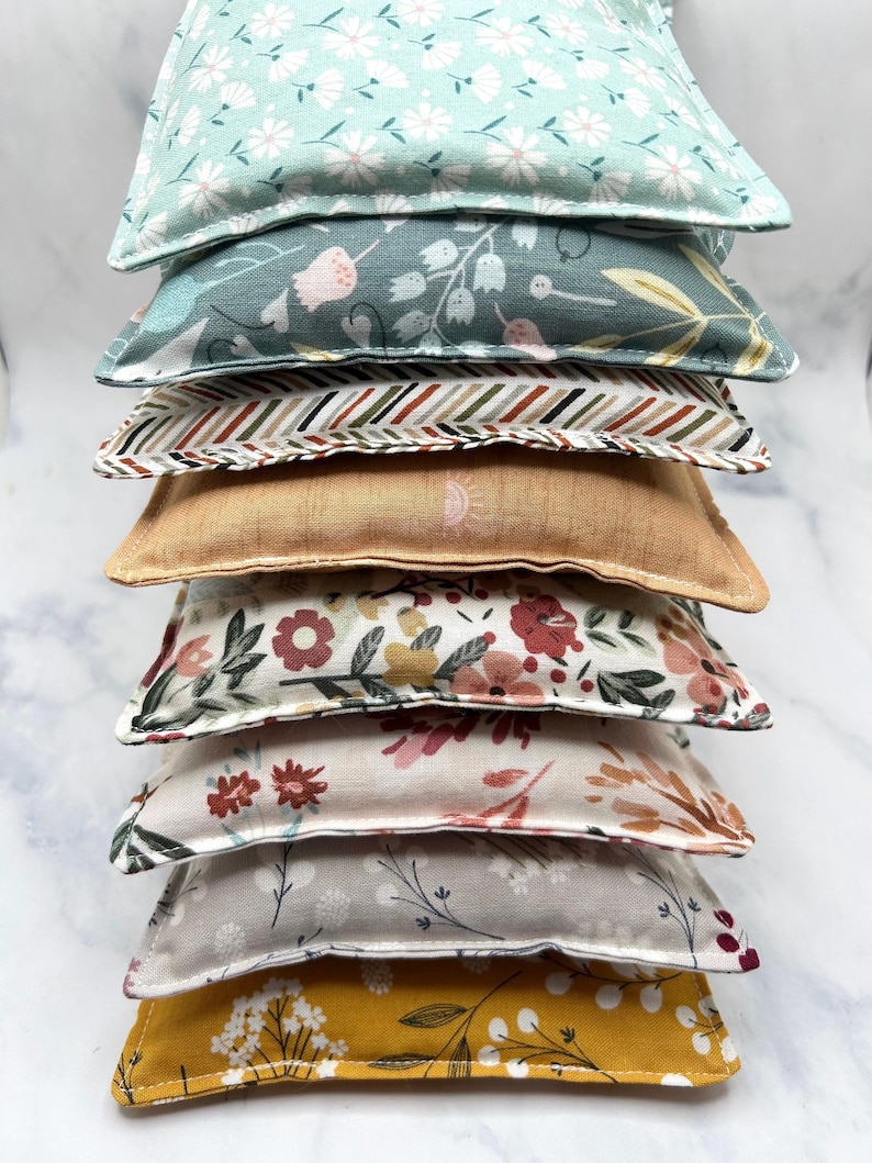May include: A stack of eight fabric-covered heat packs in various floral and geometric patterns. The heat packs are arranged from largest to smallest, with the top pack being a light blue with a white floral pattern.