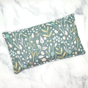 A green rectangular rice bag pillow with a floral pattern. The pattern features white, pink, and gold flowers and leaves on a green background.