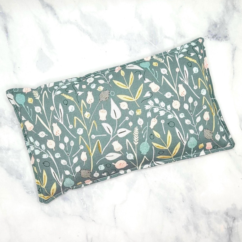 A green rectangular rice bag pillow with a floral pattern. The pattern features white, pink, and gold flowers and leaves on a green background.