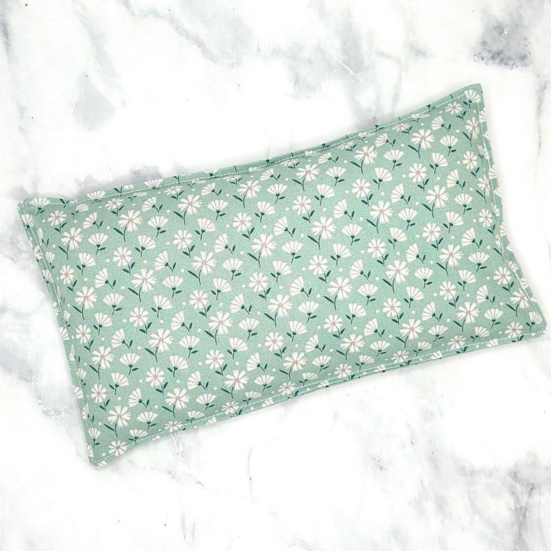 A green floral rice eye pillow with white flowers. The pillow is rectangular and has a seam around the edges.