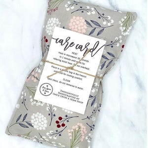 May include: A gray fabric heating pad with a floral pattern in shades of white, pink, and green. The pad is tied with twine and has a white card attached with the text "care card" and instructions for heating and cooling the pad. The card also includes the text "Thoughtfully handcrafted by Once Upon a Seam Shop."