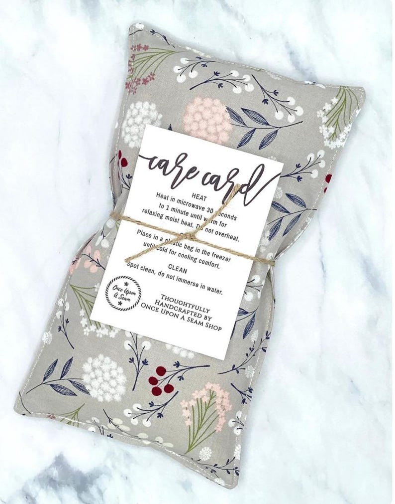 May include: A gray fabric heating pad with a floral pattern in shades of white, pink, and green. The pad is tied with twine and has a white card attached with the text "care card" and instructions for heating and cooling the pad. The card also includes the text "Thoughtfully handcrafted by Once Upon a Seam Shop."