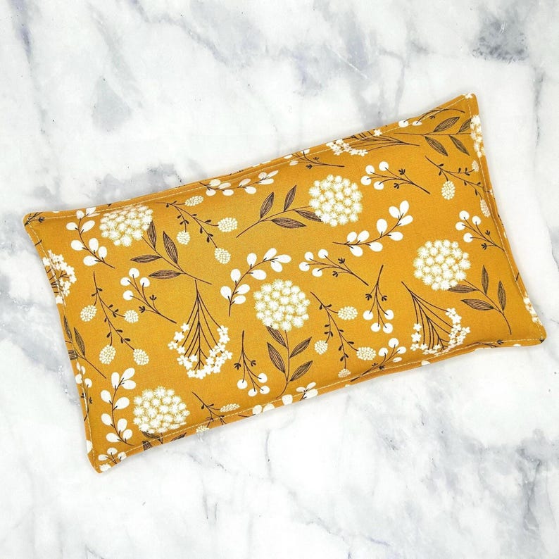 A mustard yellow floral patterned rectangular rice bag pillow with white flowers and brown stems. The pillow is made of fabric and has a soft texture.