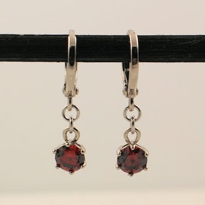 May include: A pair of silver-coloured leverback earrings. Each earring has a small chain suspending a faceted, round, deep red gemstone. The earrings are displayed against a neutral background, highlighting their design and colour.
