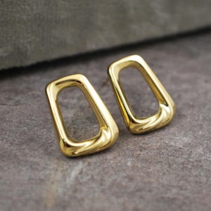 May include: A pair of gold-coloured, open-frame earrings. Each earring has a rectangular shape with rounded corners. The earrings are displayed on a grey surface, and the light reflects off the polished metal.