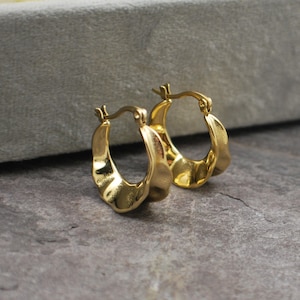 May include: A pair of gold hoop earrings. The earrings have a textured, hammered appearance and a classic hoop design. The earrings are displayed on a grey surface.