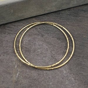 May include: Two gold bangles with a textured design are shown. The circular bangles are made of metal. The background is a dark, textured surface. The jewellery is displayed against a contrasting backdrop.