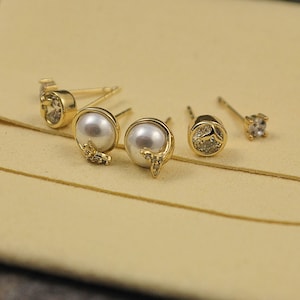 May include: Several gold stud earrings are displayed. Some have a pearl with a butterfly detail, others a clear gemstone or a circular design with a clear stone. The earrings are presented on a light tan surface.