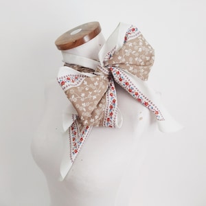 Puede incluir: Un pañuelo vintage anudado en forma de lazo. El pañuelo es blanco con un estampado floral en color beige y detalles rojos. Se muestra sobre una cabeza de maniquí.