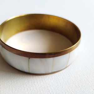Puede incluir: Un brazalete circular con un borde de metal de latón pulido en la parte superior e inferior. La sección central está compuesta por segmentos verticales rectangulares de material de concha blanca iridiscente, creando un diseño de banda lisa.
