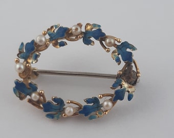 May include: A gold oval brooch with blue enamel leaves and white pearl accents.