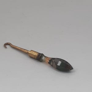 May include: A crochet hook with a brass-colored metal shaft and hook. The handle is a dark, faceted material with a gold-colored metal band near the shaft. The hook is designed for crafting.