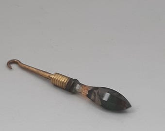 May include: A crochet hook with a brass-colored metal shaft and hook. The handle is a dark, faceted material with a gold-colored metal band near the shaft. The hook is designed for crafting.