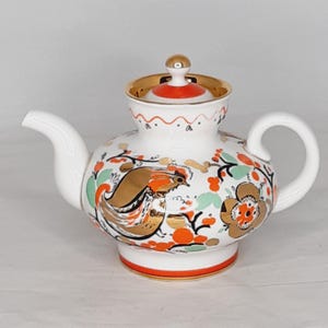 May include: White porcelain teapot with a red and gold floral design featuring a bird. The teapot has a lid with a gold knob.