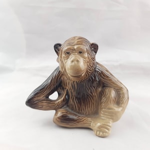 May include: A ceramic figurine of a brown and tan monkey sitting with its legs crossed. The monkey has a realistic appearance with fur details.