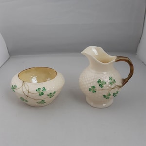 Könnte beinhalten: Cremé-farbenes Keramik-Sahne- und Zuckerdosen-Set. Der Sahneaufbereiter hat einen braunen Griff und eine strukturierte Oberfläche. Beide Teile sind mit grünen Kleeblattdesigns und braunen Stielen verziert.