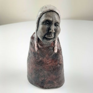 May include: A handcrafted ceramic sculpture depicting a person with a calm expression. The figure is wearing a light-coloured hat and a dark red garment. The sculpture has a textured, earthy appearance, with subtle colour and tone variations.