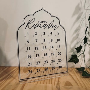 K&ouml;nnte beinhalten: Ein transparenter Ramadan-Kalender aus Acryl mit schwarzem Rahmen und dem Schriftzug "Happy Ramadan" oben. Der Kalender hat 30 nummerierte Kreise f&uuml;r jeden Tag des Monats.