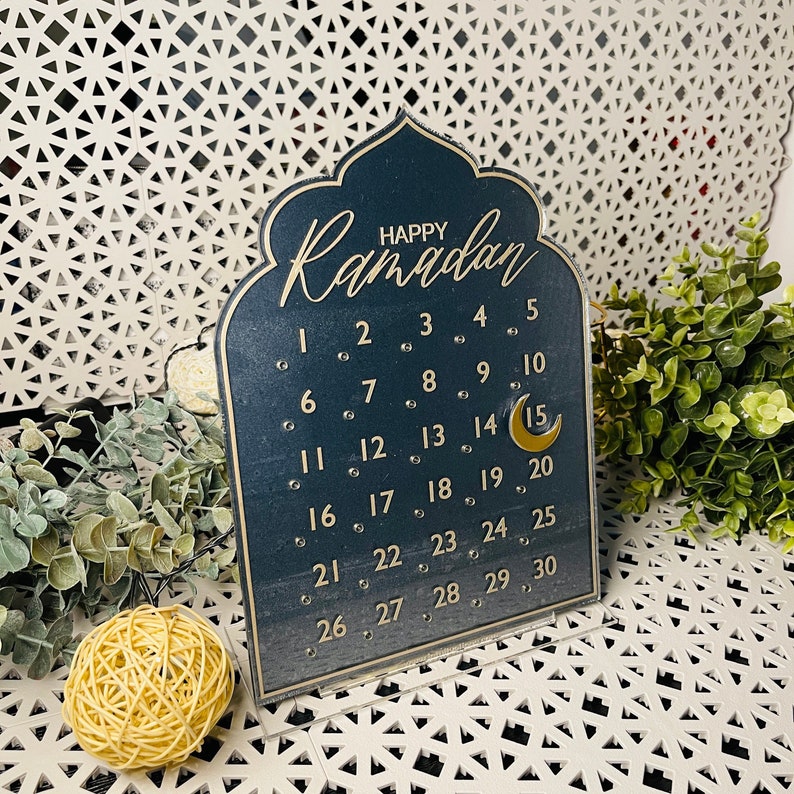 K&ouml;nnte beinhalten: Ein dunkelblauer Ramadan-Kalender aus Holz mit goldenen Buchstaben und einem Halbmond-Design. Der Kalender zeigt die Zahlen von 1 bis 30.