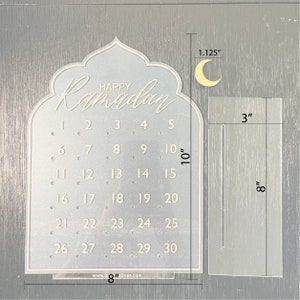 K&ouml;nnte beinhalten: Ein wei&szlig;er Ramadan-Kalender aus Acryl mit einem Halbmond-Design. Der Kalender verf&uuml;gt &uuml;ber ein Raster mit den Zahlen 1 bis 30 und den Text "Happy Ramadan" oben. Der Kalender misst 20,3 cm in der Breite und 25,4 cm in der H&ouml;he.