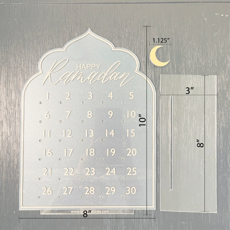 K&ouml;nnte beinhalten: Ein wei&szlig;er Ramadan-Kalender aus Acryl mit einem Halbmond-Design. Der Kalender verf&uuml;gt &uuml;ber ein Raster mit den Zahlen 1 bis 30 und den Text "Happy Ramadan" oben. Der Kalender misst 20,3 cm in der Breite und 25,4 cm in der H&ouml;he.