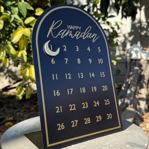 K&ouml;nnte beinhalten: Ein schwarzer und goldener Ramadan-Kalender mit einem Halbmond und den Zahlen 1 bis 30. Der Kalender hat die Form eines Bogens.