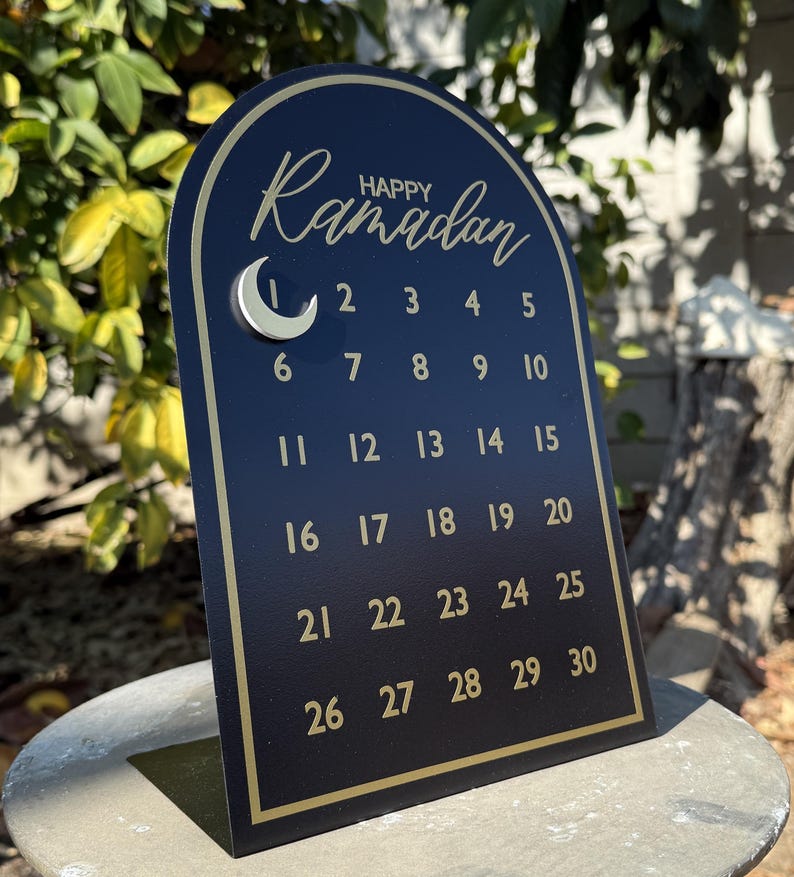 K&ouml;nnte beinhalten: Ein schwarzer und goldener Ramadan-Kalender mit einem Halbmond und den Zahlen 1 bis 30. Der Kalender hat die Form eines Bogens.