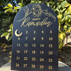 K&ouml;nnte beinhalten: Schwarzer Holzkalender mit goldener Beschriftung. Der Kalender ist bogenf&ouml;rmig und hat oben ein florales Design. Der Text "Happy Ramadan" steht oben auf dem Kalender. Die Zahlen 1 bis 30 sind in Reihen auf dem Kalender aufgelistet.